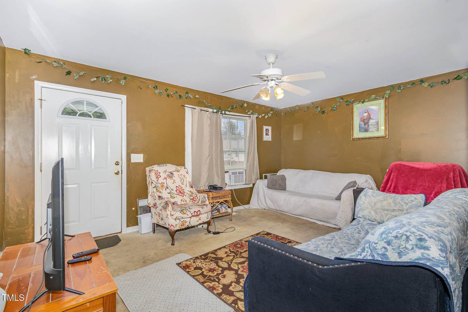 A living room with brown walls, two couches with slipcovers, an armchair, a patterned rug, a TV on a wooden stand, a ceiling fan with lights, and wall decor including a framed picture and artificial greenery.