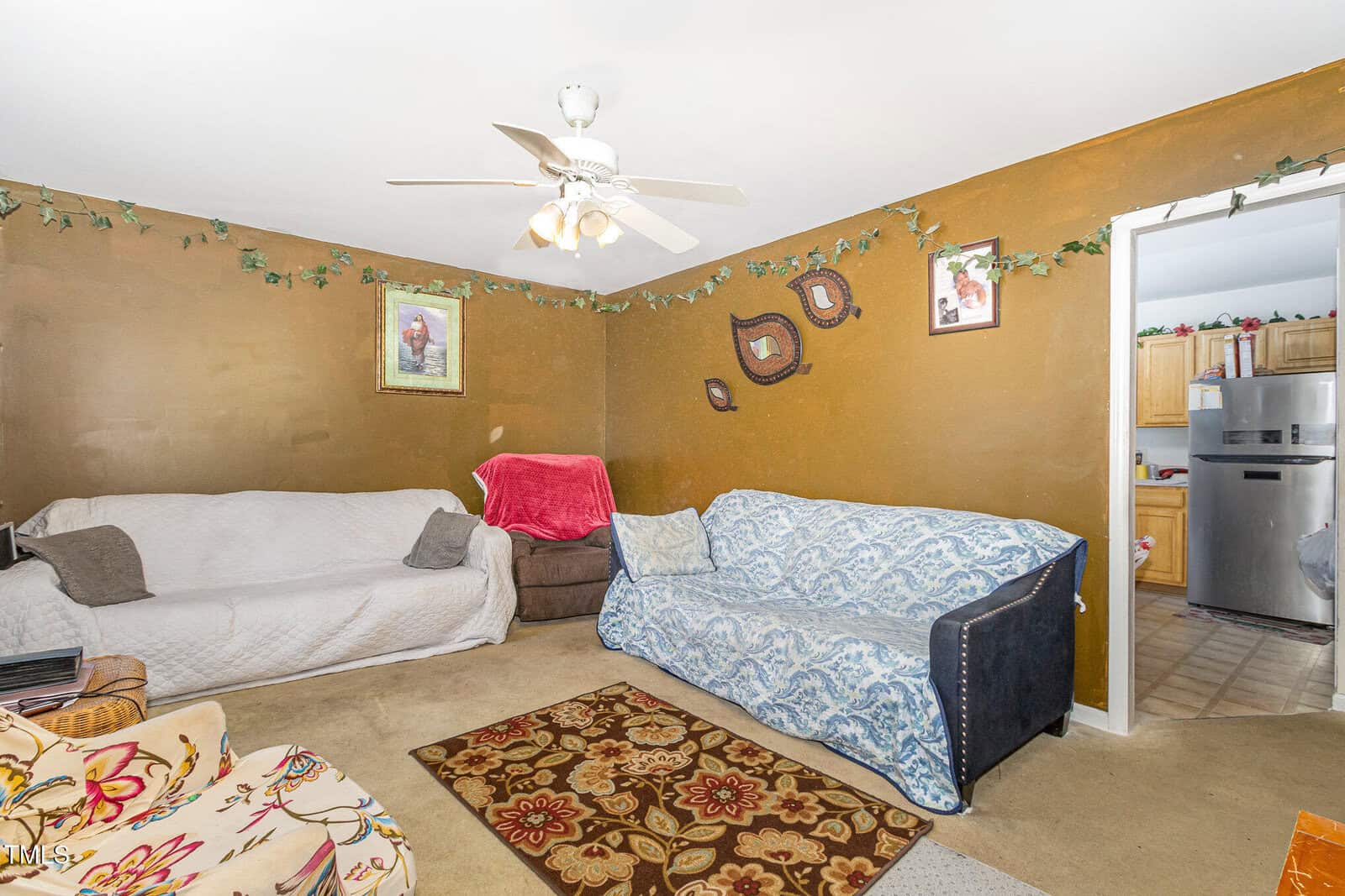 A living room with two sofas covered in slipcovers, a brown accent wall with three framed pictures, a ceiling fan with lights, a rug, and a doorway leading to a kitchen with a stainless steel refrigerator.