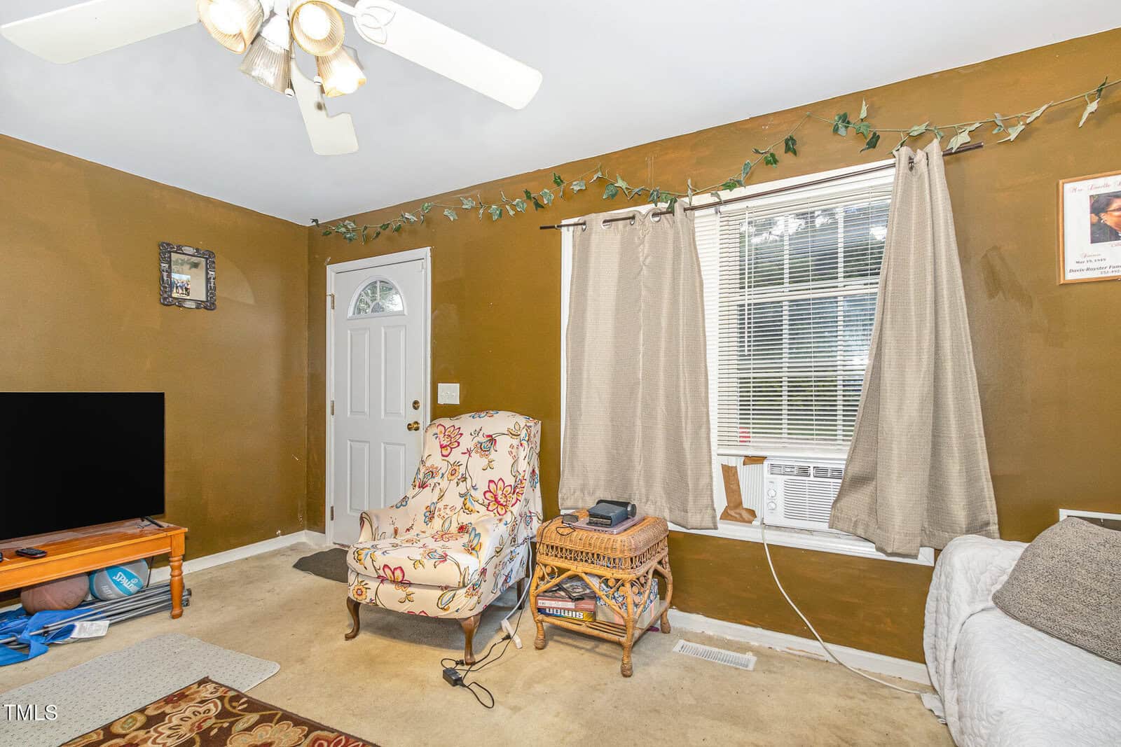 A living room with brown walls, a ceiling fan, a window with beige curtains and an air conditioner, a floral armchair, a wicker side table, a TV on a stand, and a closed white door.