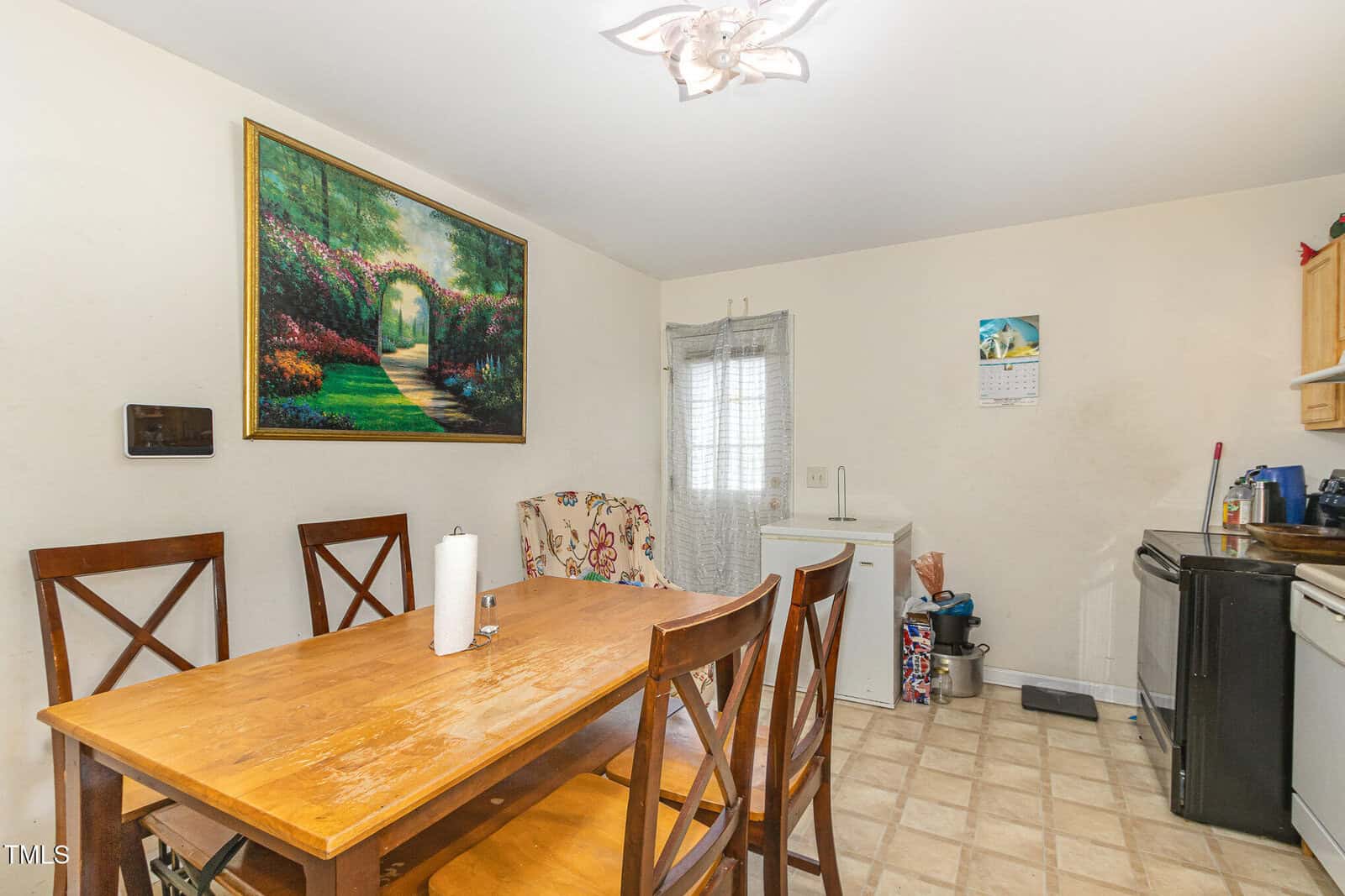 A small kitchen dining area with a wooden table and four chairs, paper towels on the table, a floral armchair, a large wall painting, a calendar, a window, a fridge, and kitchen appliances.