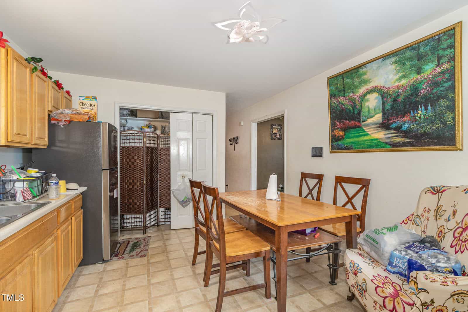 A cozy kitchen with wooden cabinets, a dining table with four chairs, a floral armchair, and a large painting of a garden path on the wall. Groceries and household items are visible on the table and chair.