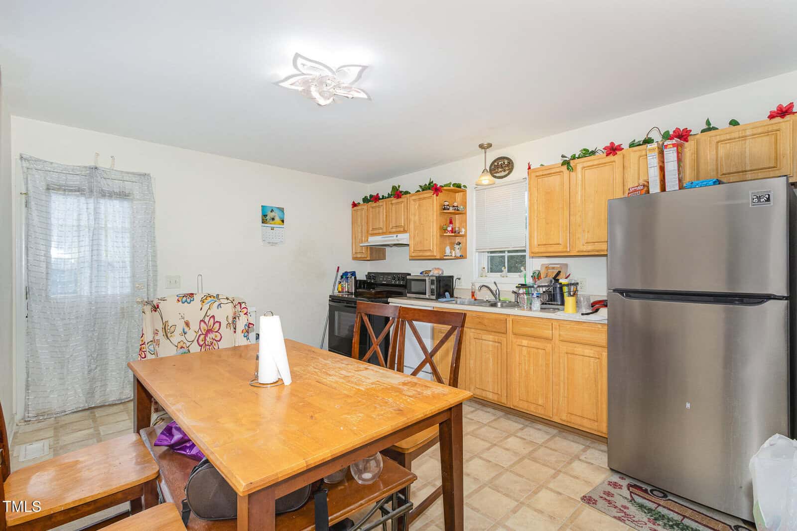A kitchen with wooden cabinets, stainless steel fridge, stove, microwave, and sink. A wooden table with chairs and paper towels is in the foreground. Decorative flowers line the cabinet tops.