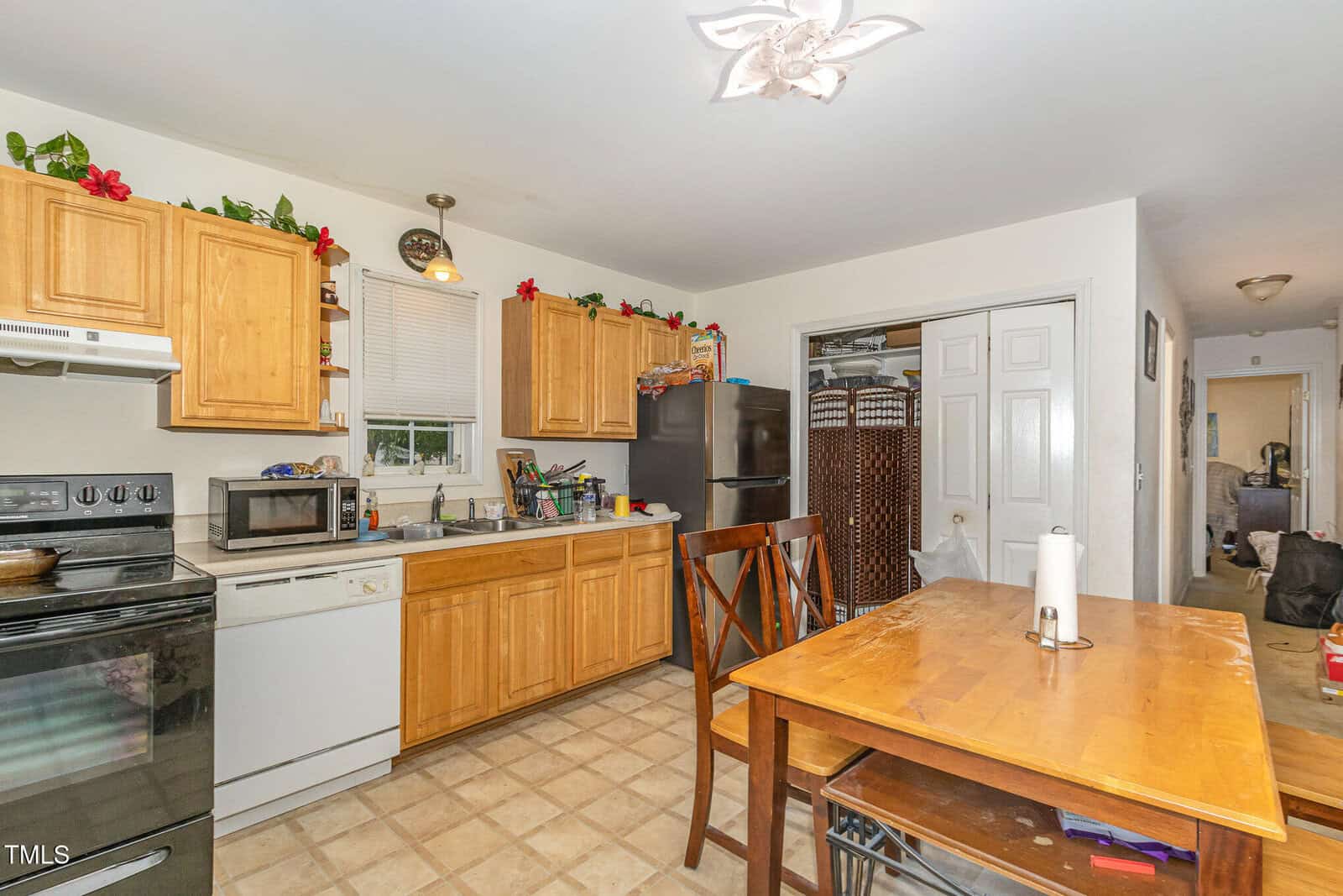 A kitchen with wooden cabinets, black appliances, and a small dining table with two chairs. There are plants and red flowers above the cabinets and various kitchen items on the counters.