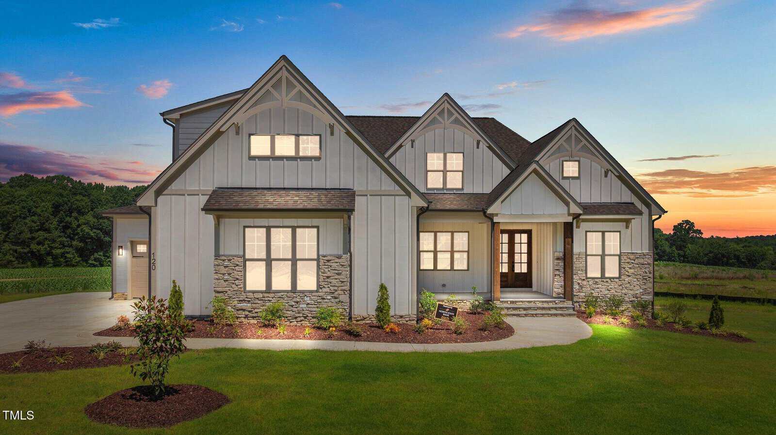 A modern two-story house with gray siding, stone accents, and large windows, illuminated at dusk. The landscaped front yard has small shrubs and a curved walkway leading to the covered entryway.