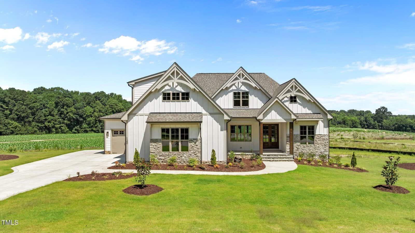 A modern farmhouse-style home with white siding, stone accents, a covered front porch, and gabled roofs, set on a large, manicured lawn with landscaped garden beds and surrounded by trees and open fields.