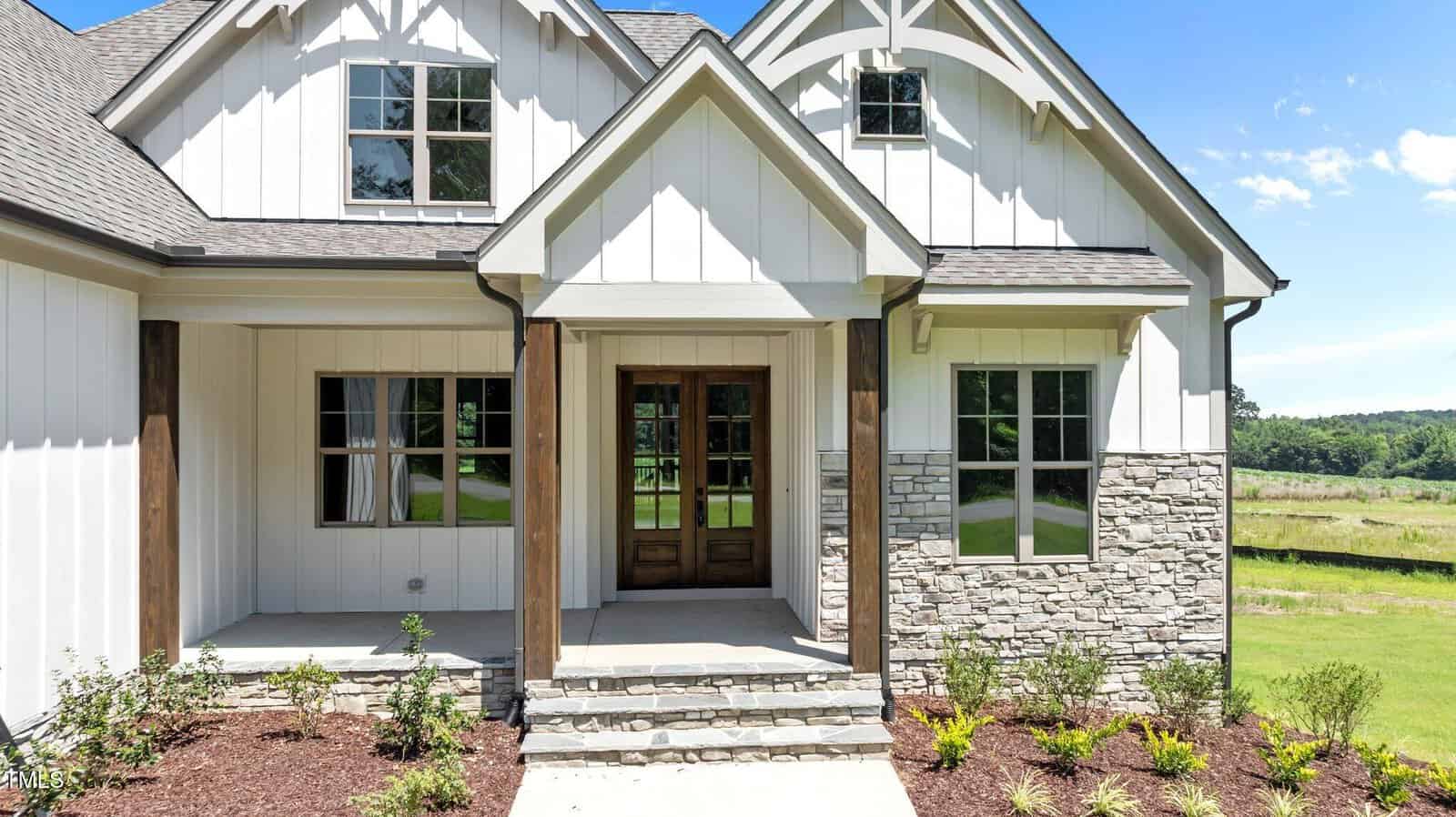 A modern farmhouse with white siding, stone accents, and wooden columns, featuring large windows and a wooden front door, surrounded by fresh landscaping and a green lawn.