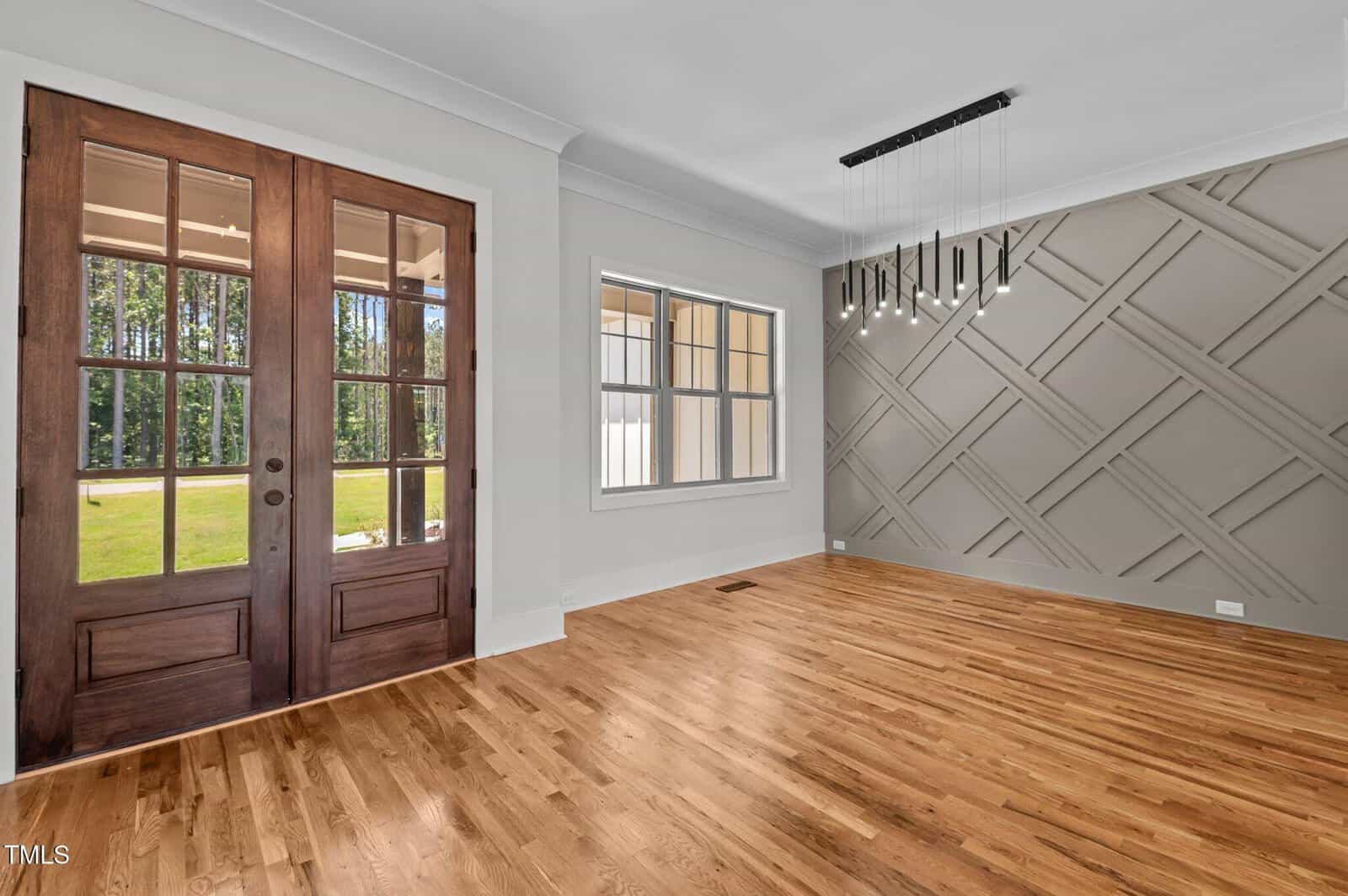 A bright, empty room with wooden floors, a modern geometric accent wall, a rectangular pendant light, double wooden doors with glass panels, and a large window overlooking a grassy yard.