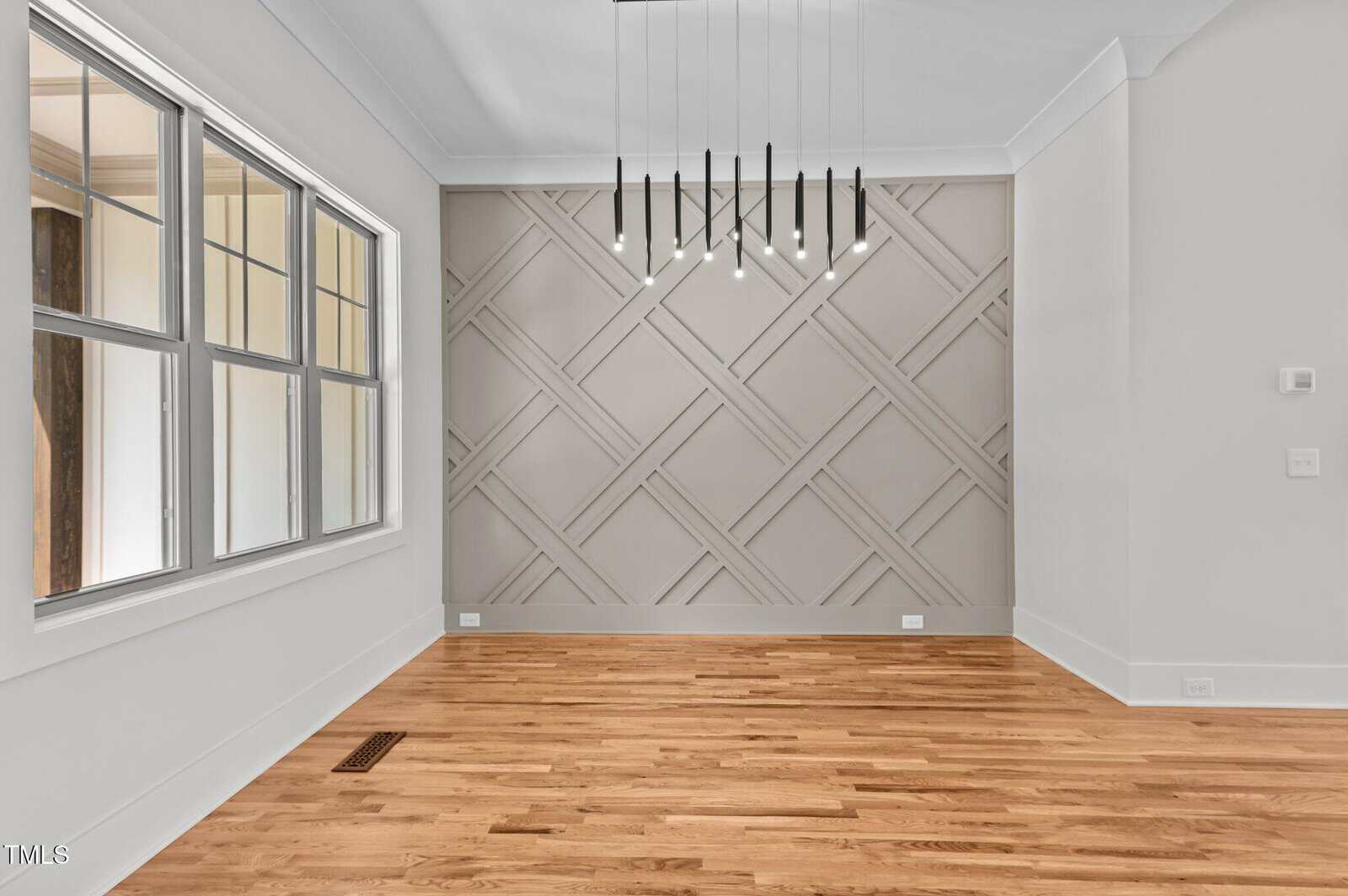 Empty room with light wood floors, geometric accent wall, multiple black pendant lights hanging from the ceiling, and large windows on the left. Walls and trim are painted white. No furniture is present.