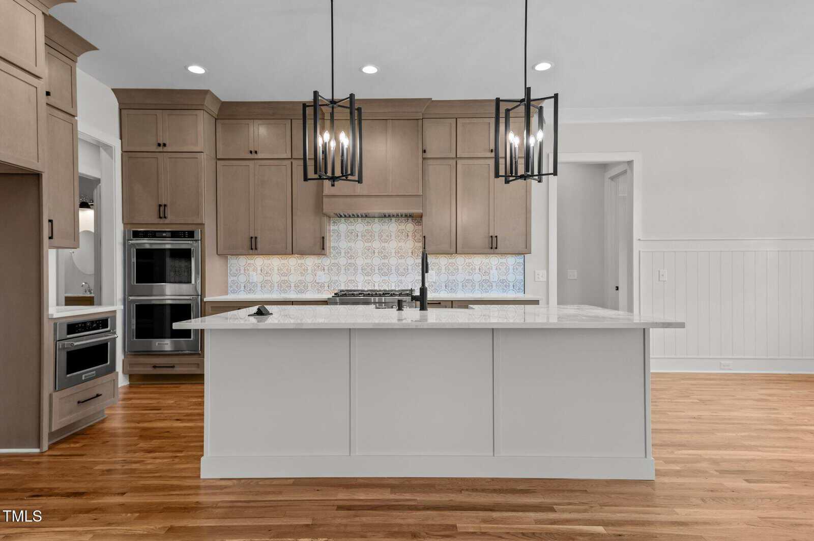 Modern kitchen featuring a large white island with marble countertop, wood cabinets, double wall ovens, a gas cooktop, patterned tile backsplash, pendant lights, and hardwood floors.