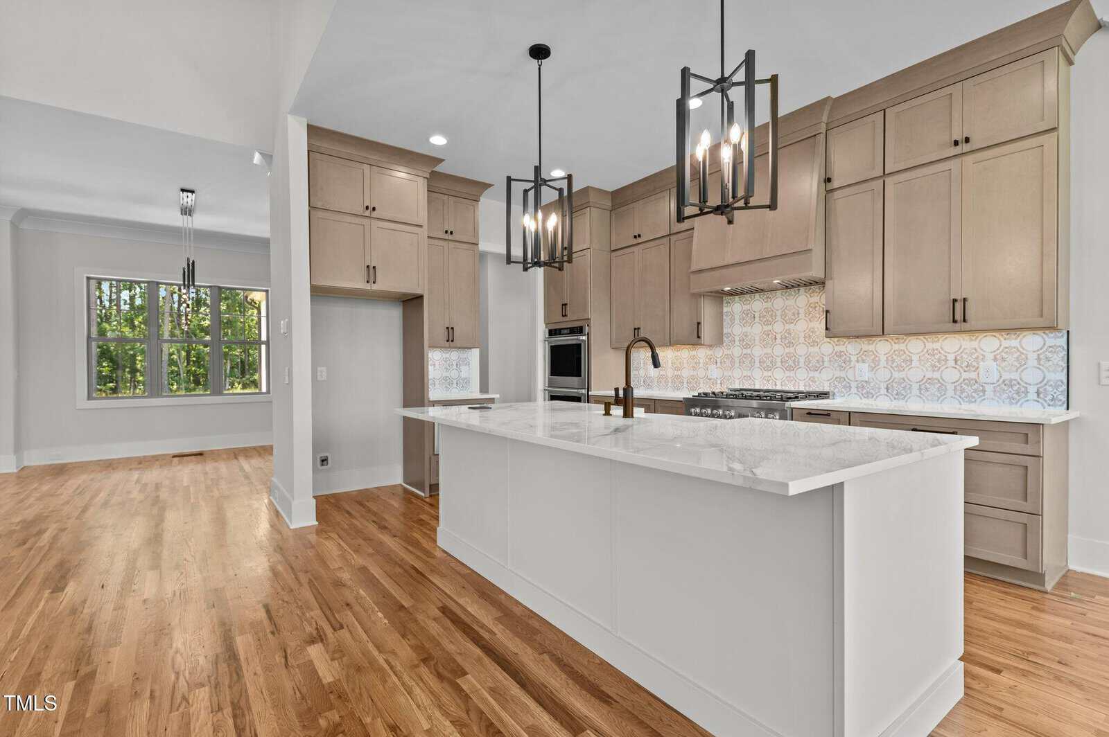 Modern kitchen with beige cabinets, white marble island, patterned tile backsplash, stainless steel appliances, and two pendant lights. Hardwood floors extend into an adjacent dining area with a large window.