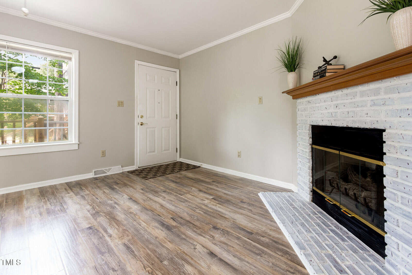 A bright living room with light gray walls, wood-look flooring, a white front door, a window letting in daylight, and a brick fireplace with a wooden mantel decorated with potted plants.