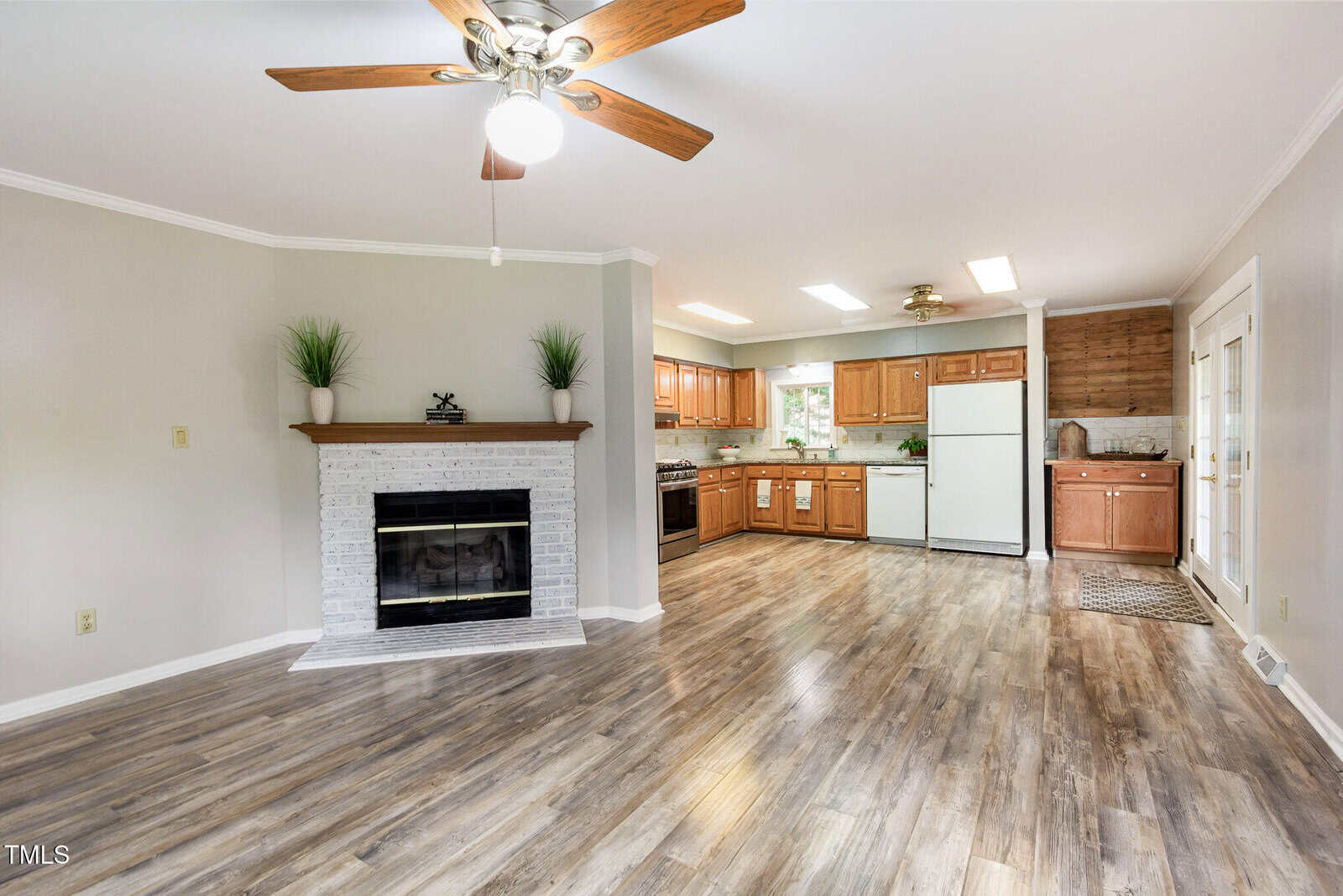Open-concept living room and kitchen with wood flooring, a white brick fireplace, ceiling fan, wooden cabinets, white appliances, and double glass doors leading outside. Bright and spacious with neutral colors.