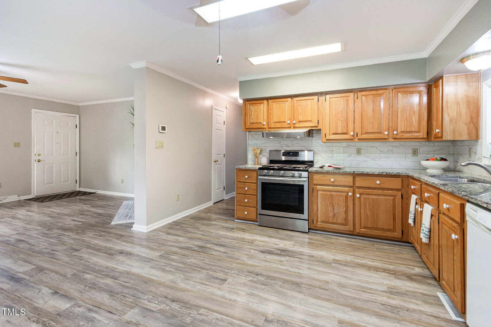 Modern kitchen with wooden cabinets, stainless steel oven, granite countertops, marble backsplash, and wood-look flooring. Open layout leads to a neutral-toned entryway with a white door and ceiling fan.