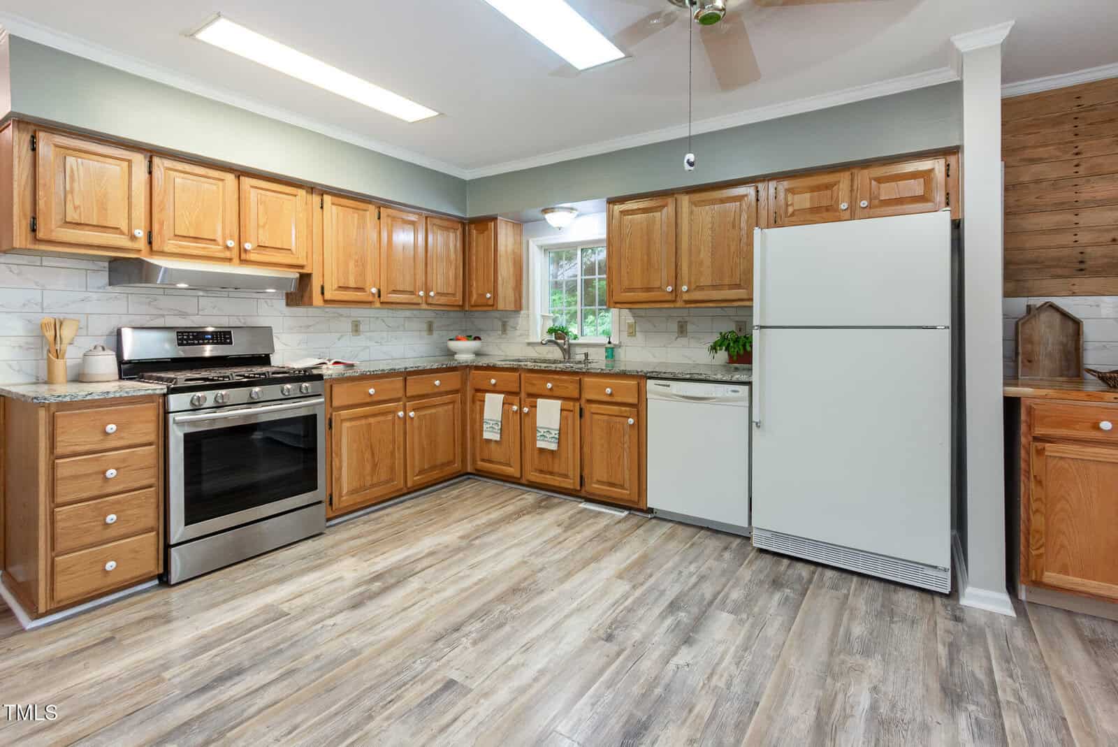 A kitchen with wood cabinets, stainless steel gas stove, white refrigerator, dishwasher, and window over the sink. The floor is light wood, and countertops are light-colored with a white tile backsplash.