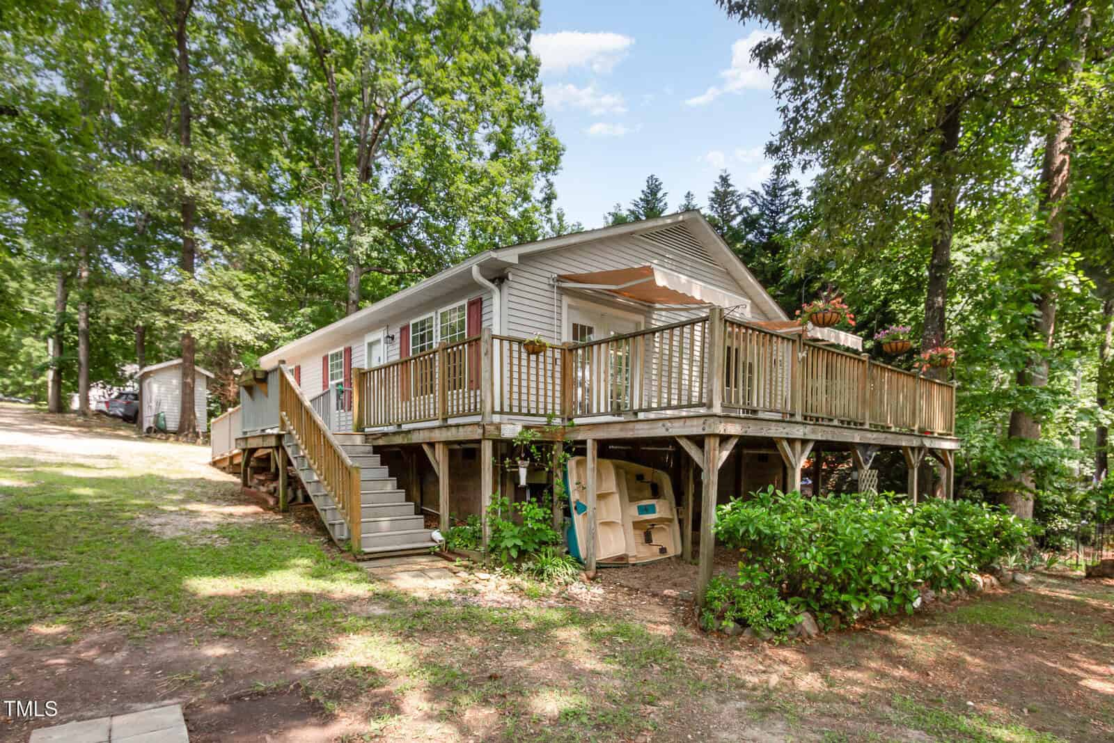 A light-colored house with a raised wooden deck, stairs leading up, and greenery around. The house sits among tall trees with a grassy yard and a small shed in the background.