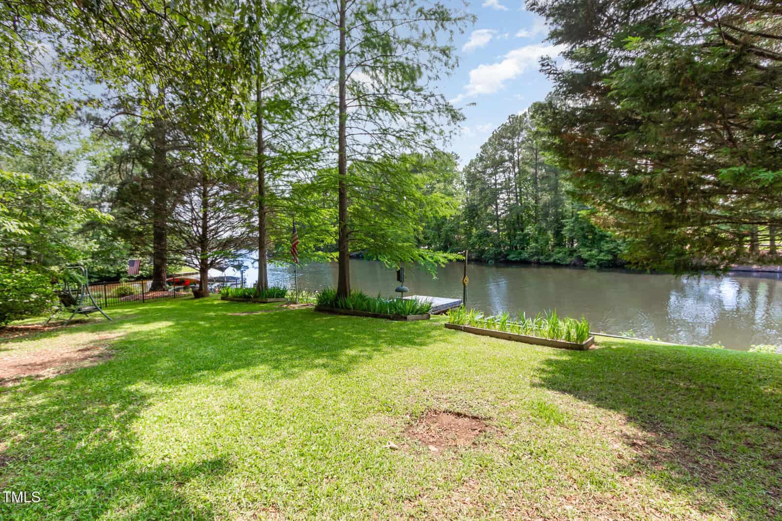 A grassy backyard overlooks a calm river, bordered by tall trees. A small dock with a bench extends over the water. Shadows from the trees create a peaceful, shaded area on a sunny day.