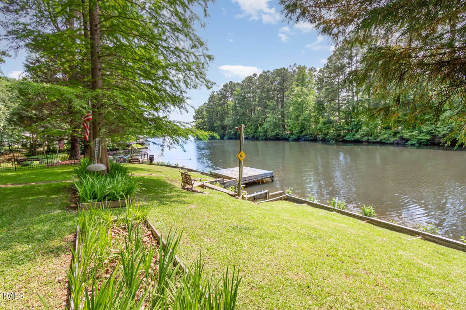 A grassy backyard slopes down to a calm river with a small wooden dock. Tall trees line the water and provide shade, while a bench and garden beds add to the peaceful, natural setting.