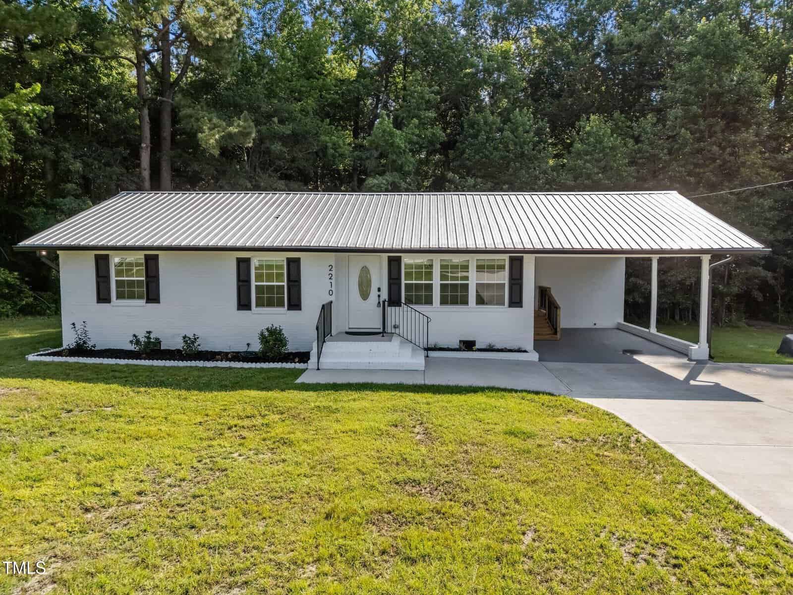 A single-story white house with black shutters, a metal roof, front steps with railings, a small garden bed, and an attached carport, set against a backdrop of trees and green lawn.