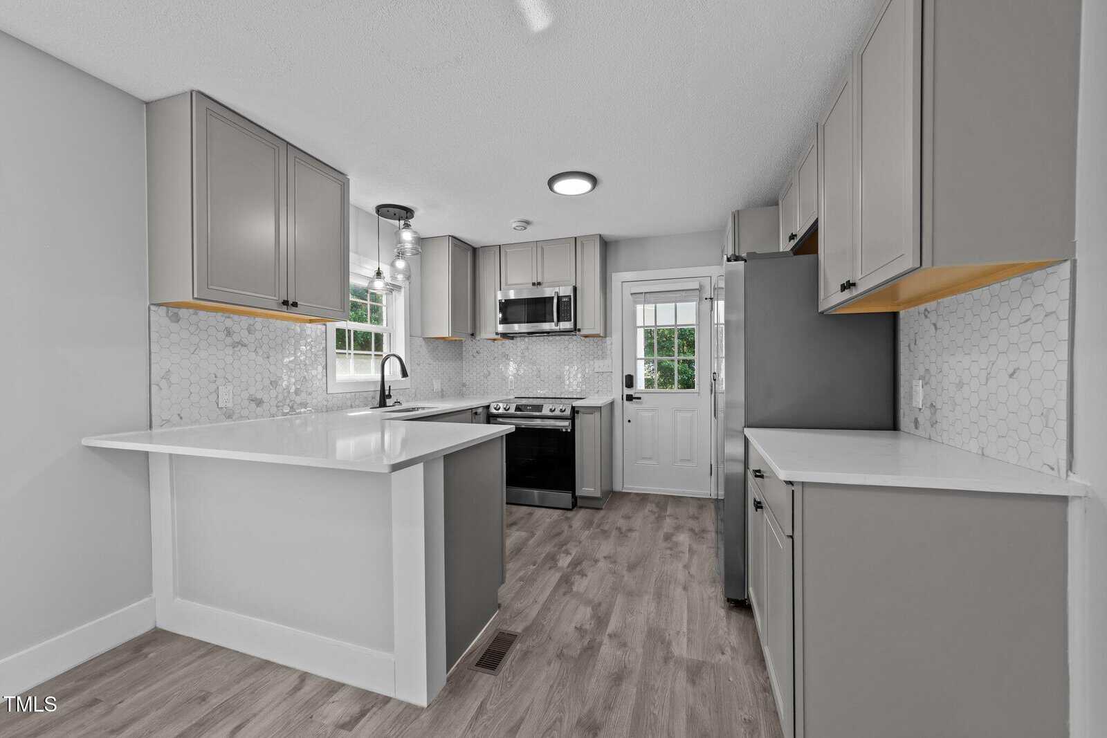 Modern kitchen with gray cabinets, white countertops, hex tile backsplash, stainless steel appliances, wood-look flooring, a window above the sink, and a door leading outside. The space is clean and well-lit.