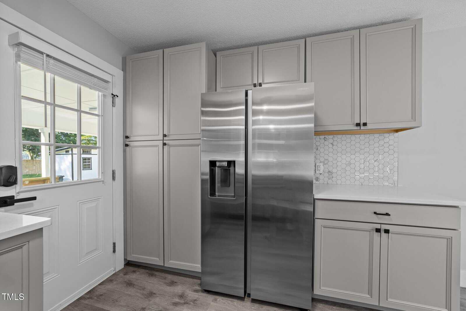 A modern kitchen features a stainless steel side-by-side refrigerator, light gray cabinets, a white hexagon tile backsplash, wood flooring, and a windowed back door with a view of the outdoors.