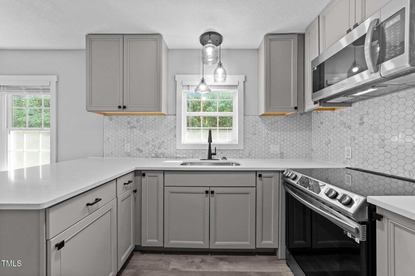 Modern kitchen with light gray cabinets, white countertops, hexagonal tile backsplash, stainless steel stove and microwave, a window above the sink, and pendant lighting overhead.