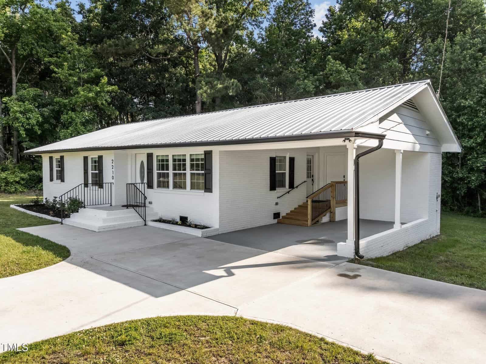 Single-story white brick house with a metal roof, black shutters, front porch, and a carport on the right. Surrounded by trees, the home has a concrete driveway and steps leading to the front door.