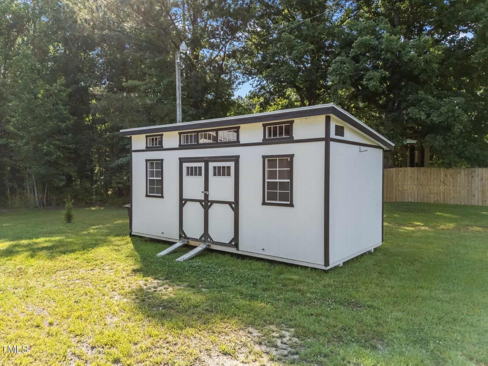 A small white shed with dark trim, double doors, and windows sits on a grassy lawn near a wooded area and a wooden fence under a sunny sky.