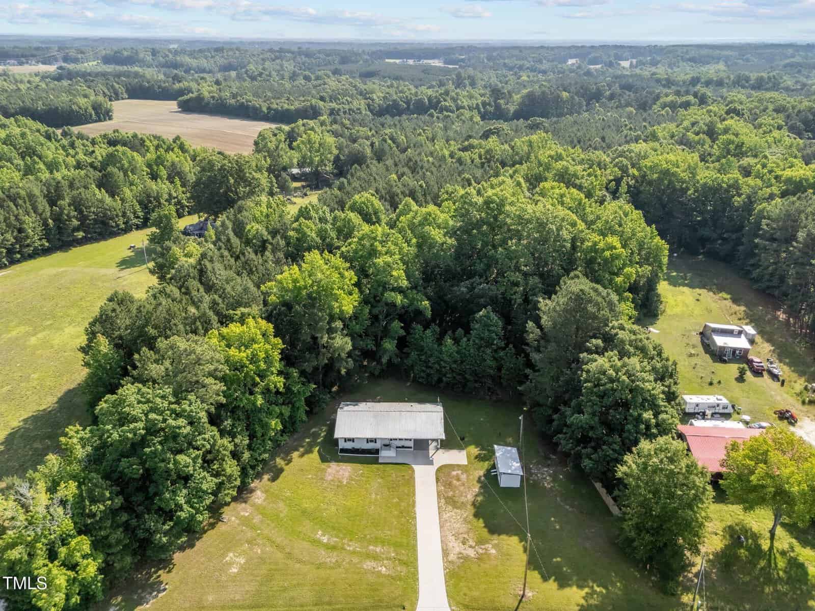 Aerial view of a white house with a driveway, surrounded by green lawns, dense trees, and forested land under a partly cloudy sky. Outbuildings and open fields are visible in the background.