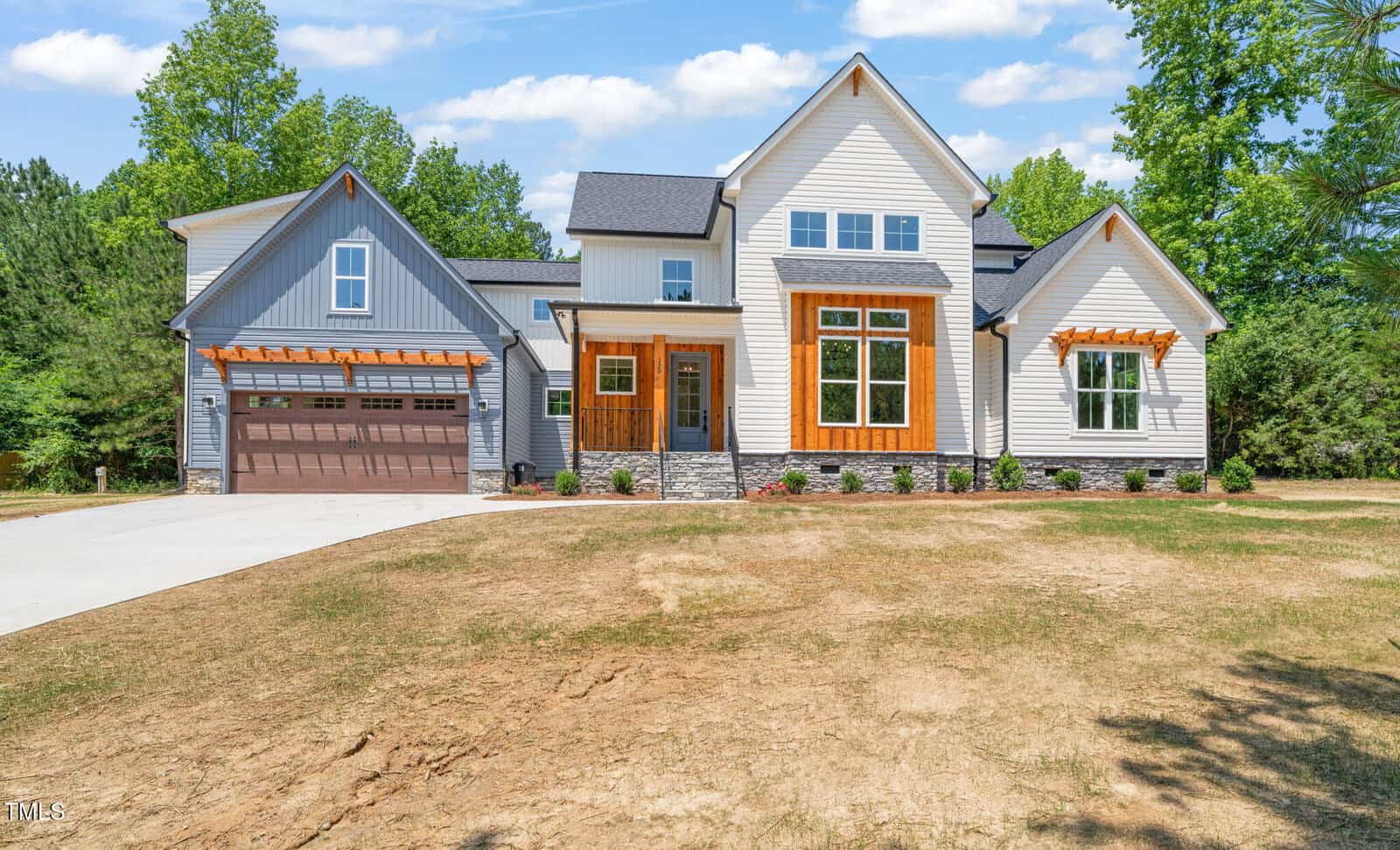 Modern two-story house with light gray siding, large windows, wooden accents, and a stone foundation. There is a double garage with a wooden door, a wide driveway, and minimal landscaping in the front yard.