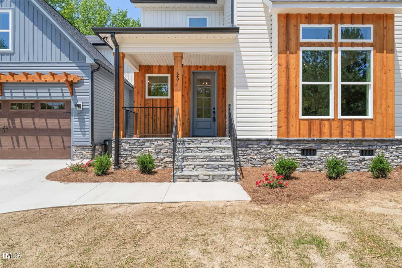 A modern house exterior with white siding, stone steps, a gray front door, wooden accents, and landscaped shrubs along the walkway. A garage with brown doors is partially visible on the left.