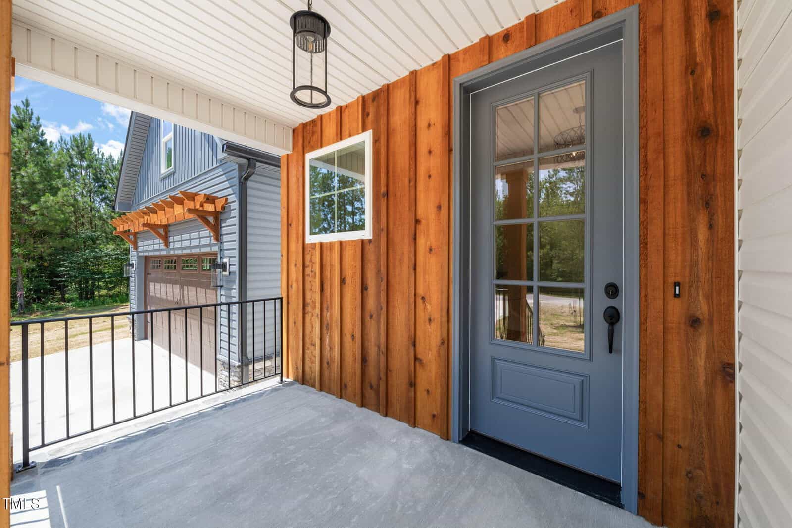A covered porch with a light fixture, featuring a wooden wall and a blue-gray glass-paneled door. Black metal railing lines the porch, and a driveway and garage with wooden accents are visible outside.