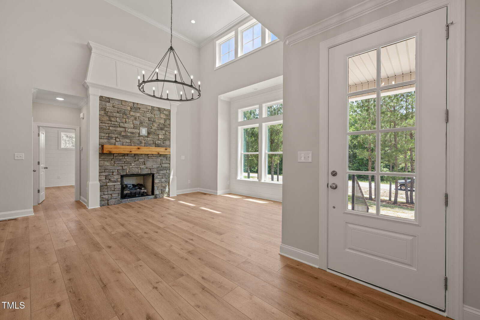 Bright, modern living room with large windows, a glass door, light wood flooring, and a stone fireplace with a wooden mantle. Sunlight fills the space, highlighting high ceilings and a contemporary chandelier.