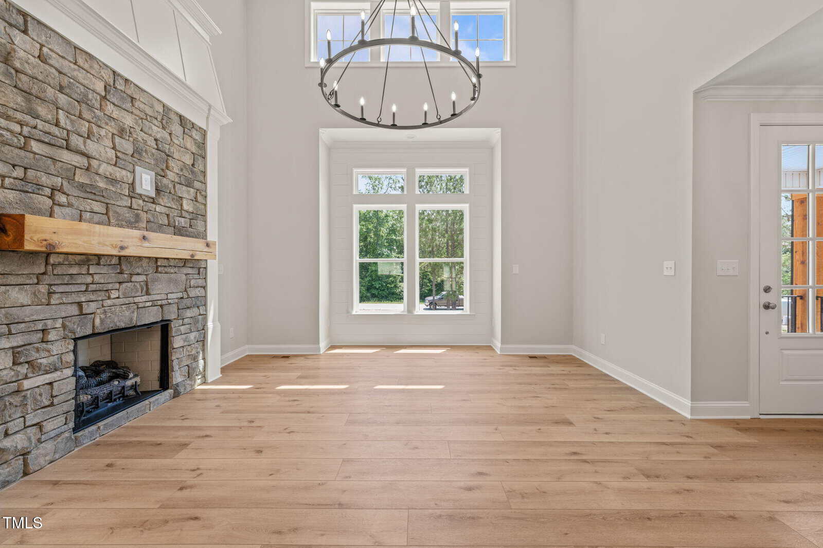 A bright, empty living room with tall ceilings, large windows letting in natural light, a stone fireplace with a wooden mantel, and light wood flooring. A modern chandelier hangs from the ceiling.