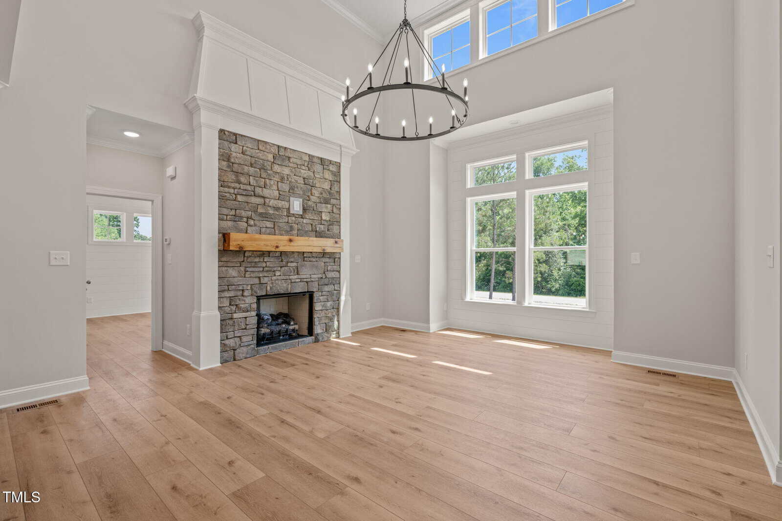Bright living room with large windows, light wood flooring, a stone fireplace with a wood mantel, high ceiling, and a round metal chandelier. White walls and wood trim are visible, with an open doorway leading to another room.