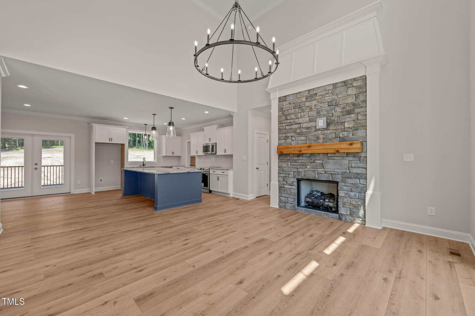 Spacious open-concept living area with light wood floors, a stone fireplace with a wood mantel, a large chandelier, and a kitchen featuring a blue island and white cabinets. Natural light enters through glass doors.