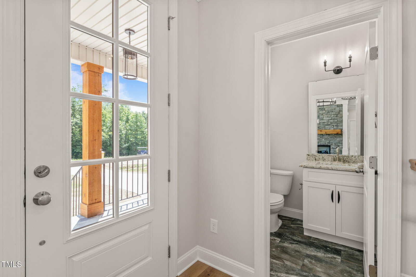 A white front door with glass panes opens to an entryway with a view of a small bathroom featuring a toilet, granite countertop, white vanity, mirror, and wall sconce lighting. Outdoor porch with wood columns is visible.