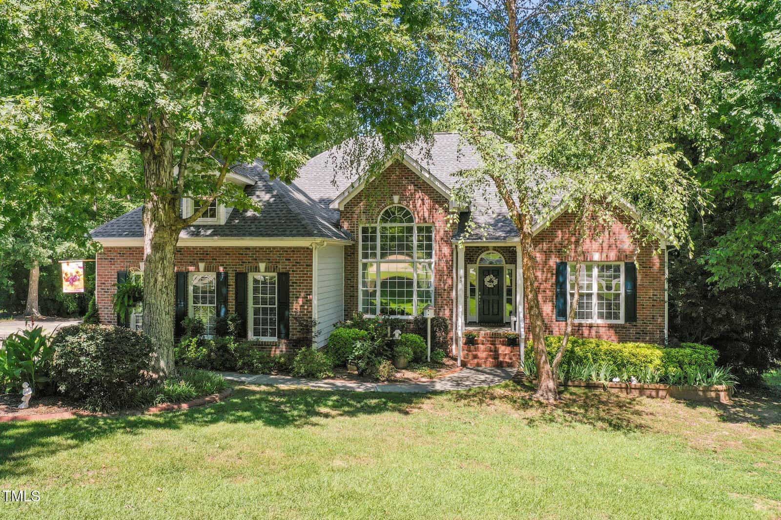 A brick and siding house with large arched windows, a dark front door, and a well-kept front yard with green grass, shrubs, and mature trees casting shade over the property.