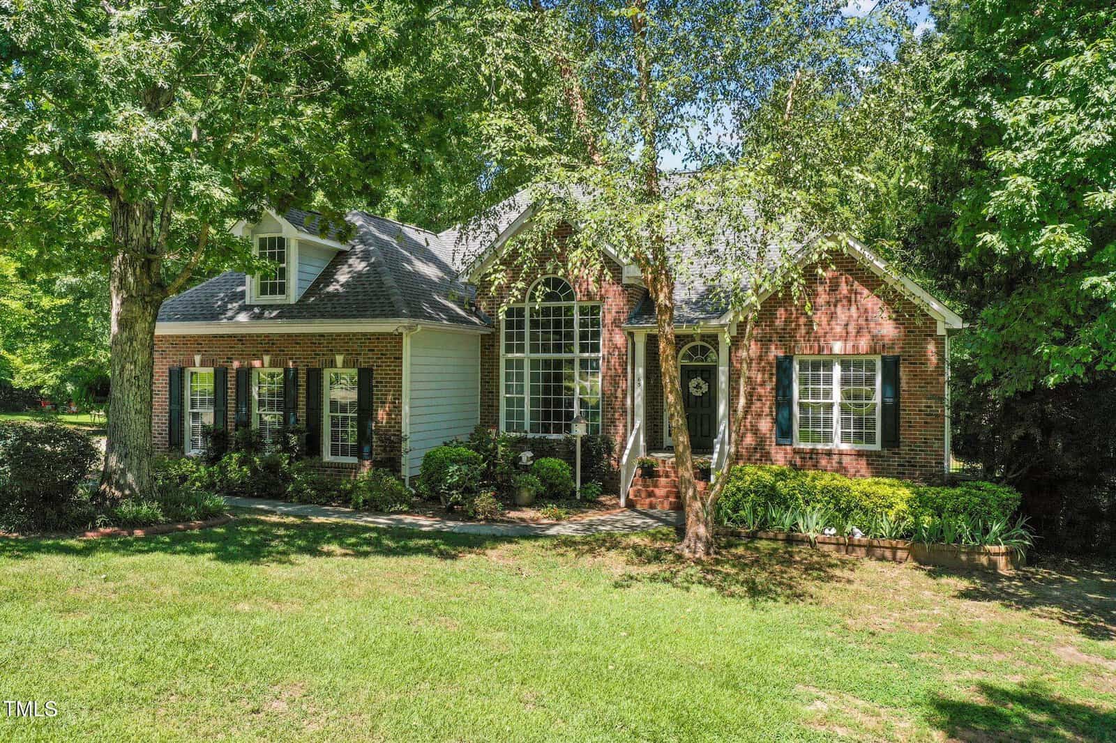 A charming brick and siding house with large arched windows, surrounded by lush green grass, mature trees, and landscaping on a sunny day.