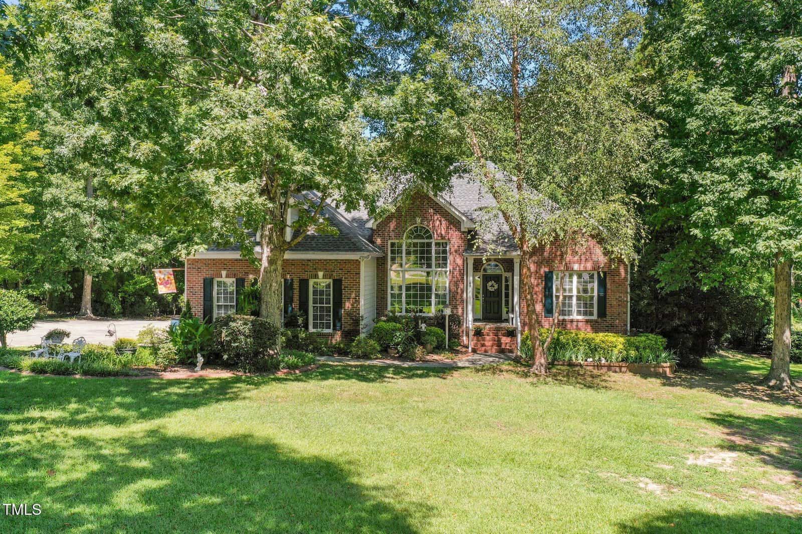 A brick house with large windows sits among tall trees and lush green grass, with a driveway to the left and abundant landscaping in the front yard.