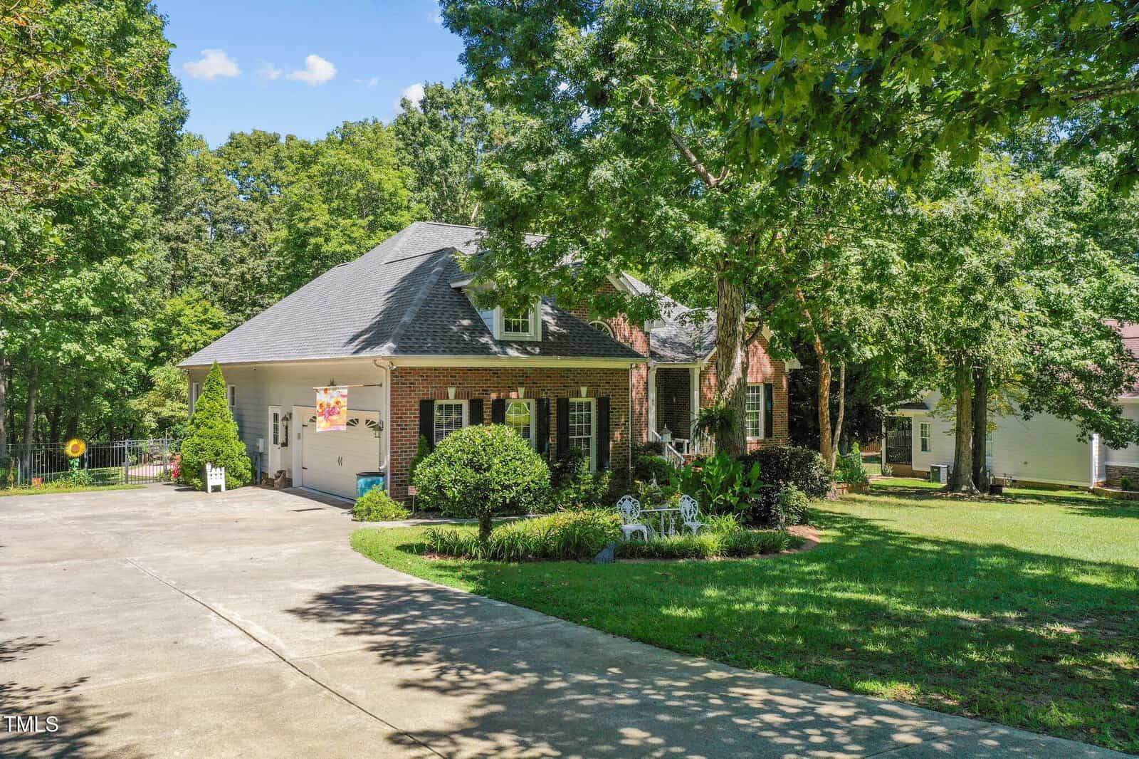 A brick house with a gray roof sits among tall trees, featuring a front porch with white chairs, a landscaped yard, and a wide driveway curving to a two-car garage.