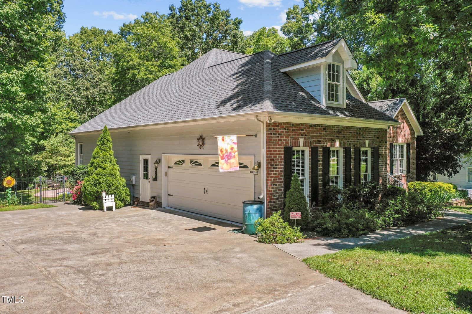 A two-story brick and siding house with a gabled roof, attached garage, and dormer window. A colorful flag hangs near the garage, and the driveway is surrounded by green trees, bushes, and a manicured lawn.