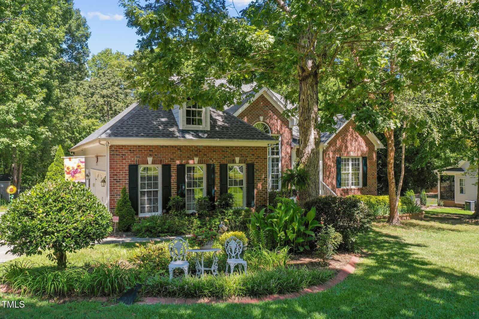 A brick house with black shutters and white trim sits in a landscaped yard with green grass, shrubs, and tall trees. A small white metal table and two chairs are on the lawn in front of the house.