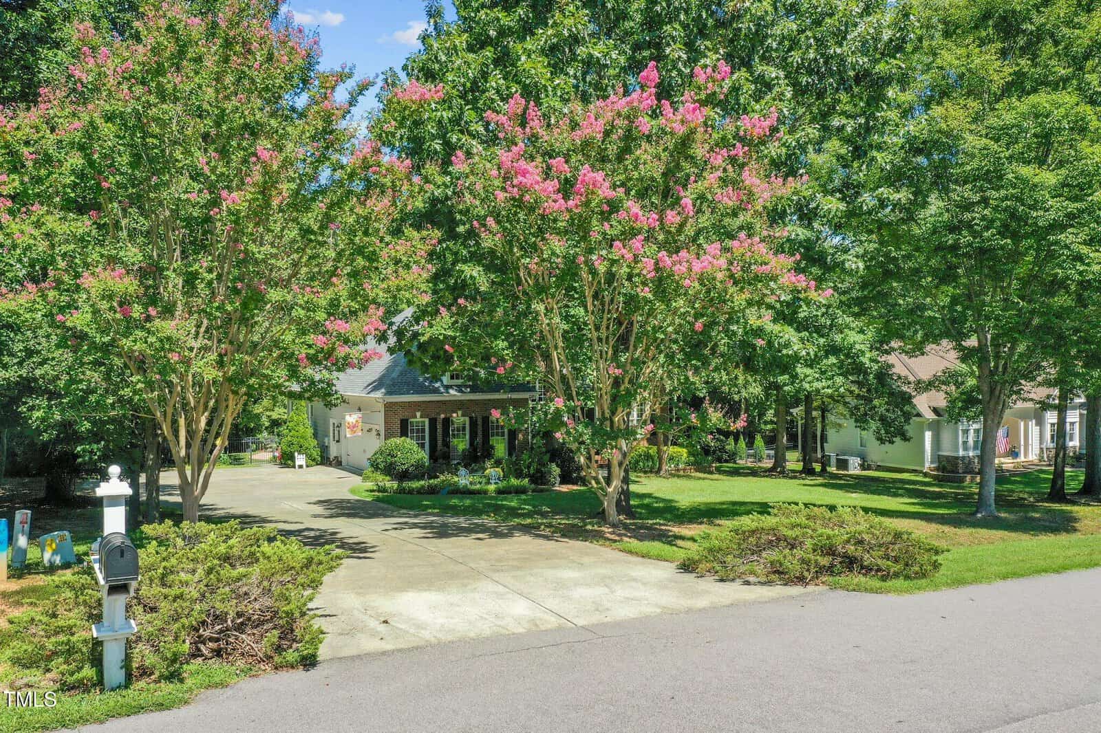 Single-story house with a covered porch, surrounded by mature trees and flowering pink crepe myrtles. Concrete driveway leads to a garage. Mailbox and street are visible in the foreground under a clear blue sky.