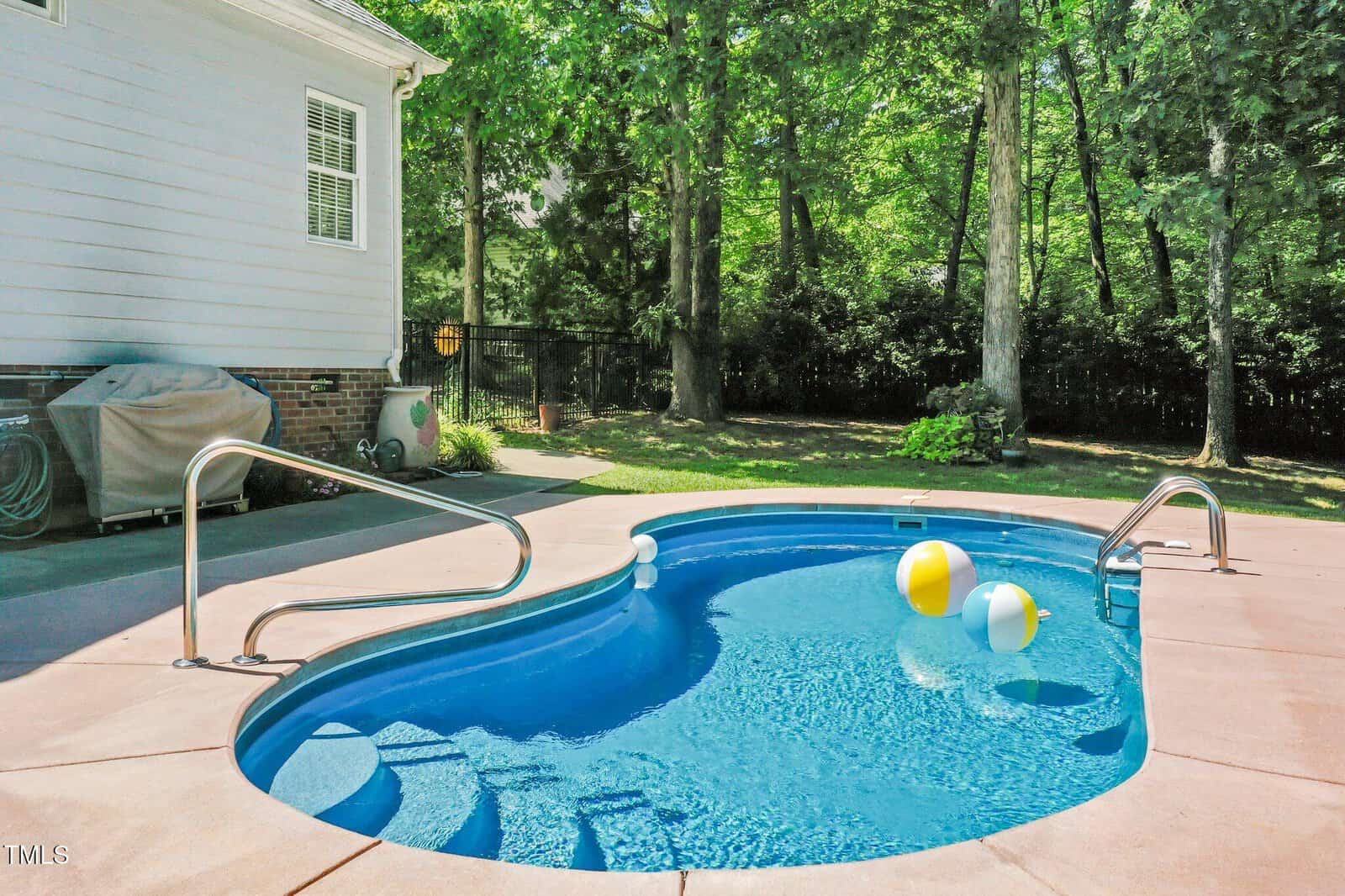 Small kidney-shaped backyard pool with clear blue water, three beach balls floating, metal handrails, and a grill covered with a tarp on the left. Surrounded by trees and a patio area on a sunny day.