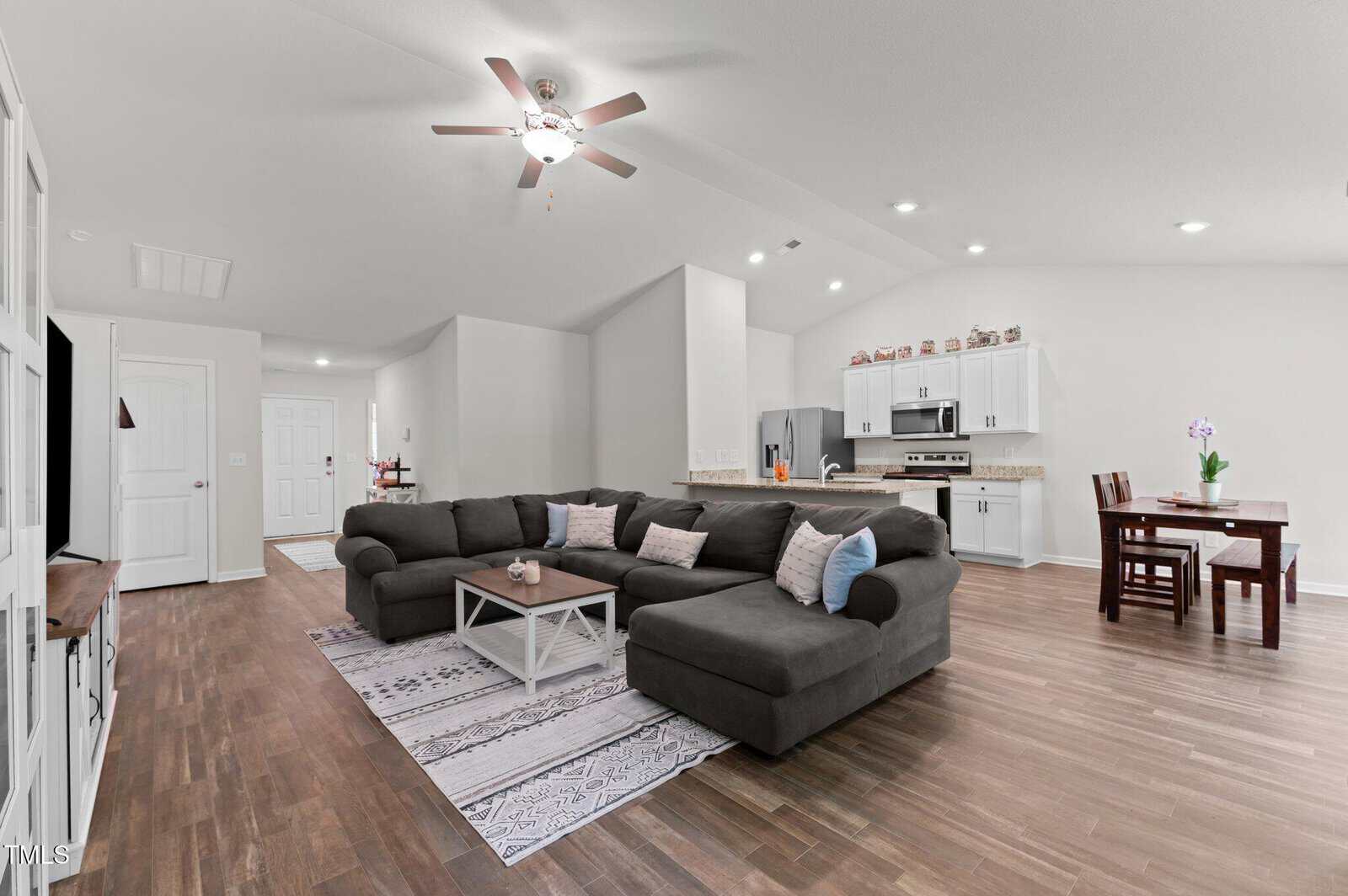 Open-concept living area with a gray sectional sofa, coffee table, rug, white walls, wood flooring, kitchen in the background with stainless steel appliances, and a dining table with four chairs. Ceiling fan and recessed lighting above.