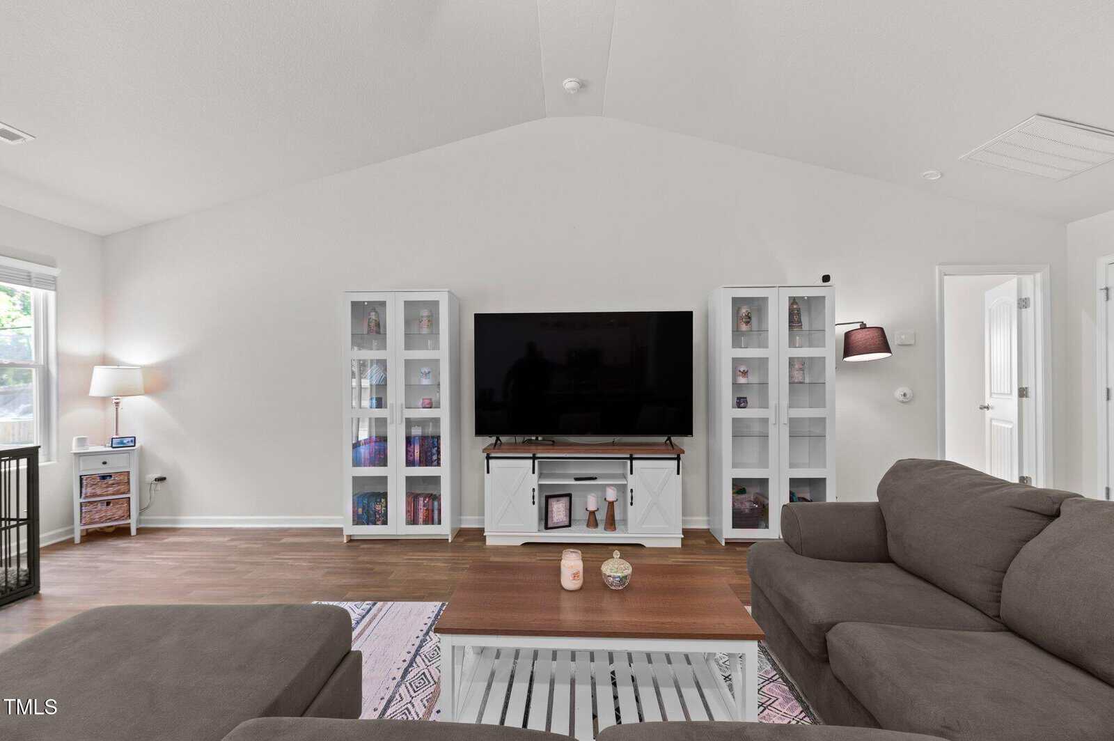 A living room with a large flat-screen TV on a white stand, flanked by two glass-door cabinets. There is a brown sectional sofa, a wooden coffee table, a side table with a lamp, and hardwood floors.