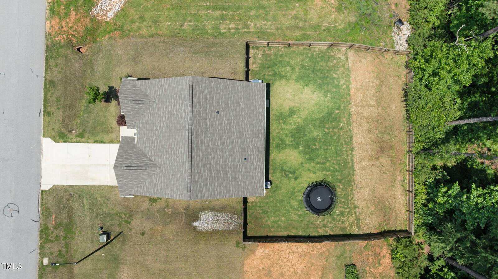 Aerial view of a suburban house with a gray roof, driveway, and fenced backyard containing a trampoline. The yard has patches of green and brown grass, and the property is bordered by a street and trees.
