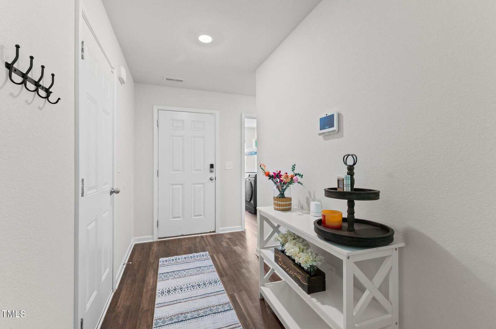 Bright entryway with white walls and a dark wood floor, featuring a striped rug, a console table with decor items, a wall hook, and a closed white door at the end of the hallway. A laundry room is visible to the right.