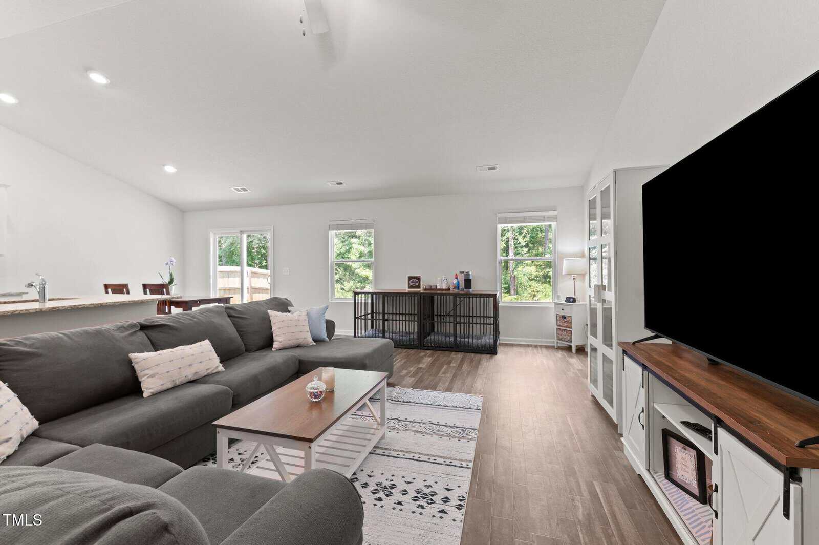 A modern living room with a large gray sectional sofa, white coffee table, patterned rug, TV on a white stand, hardwood floors, and three windows letting in natural light. The kitchen and dining area are visible in the background.