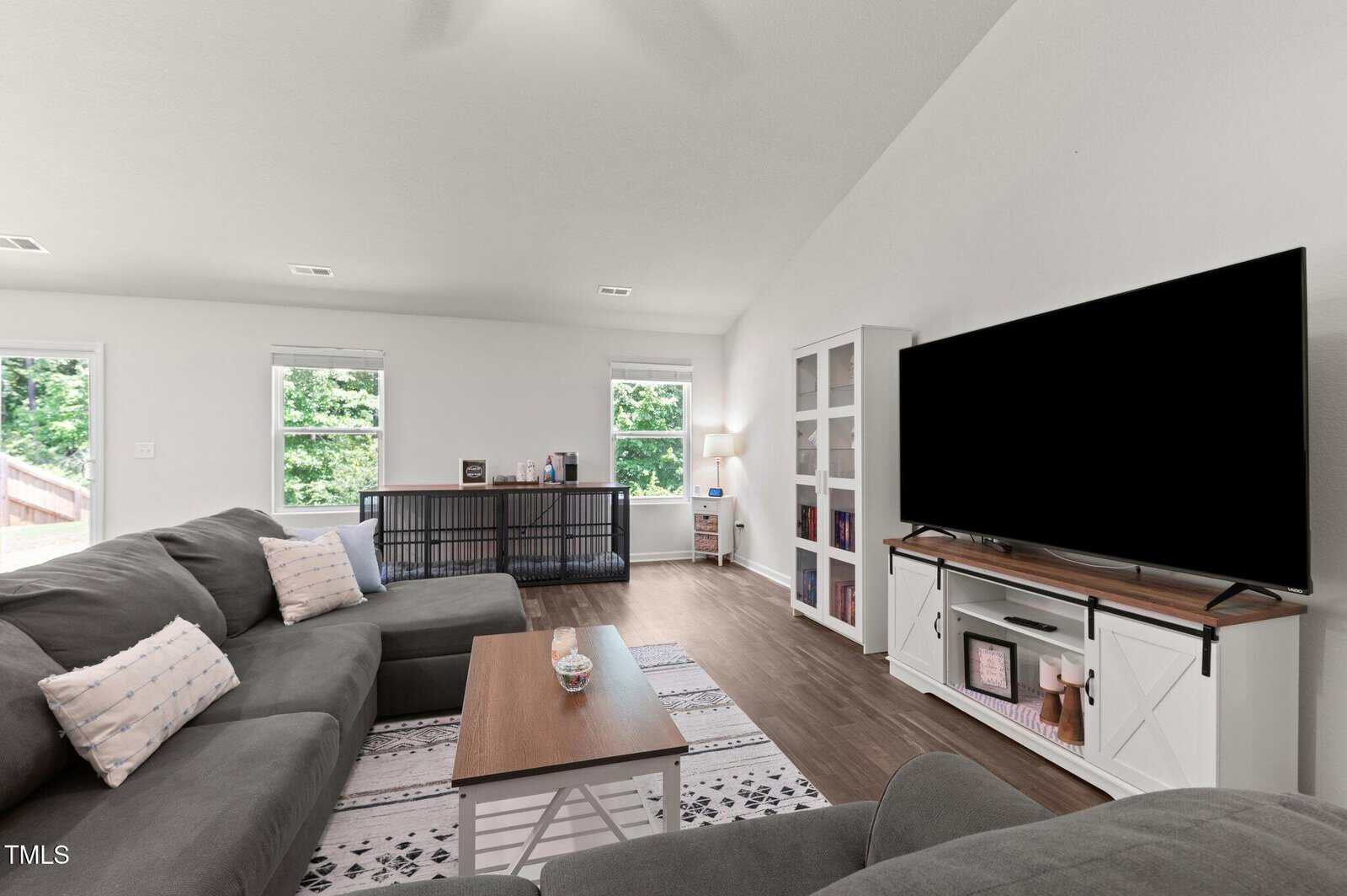 Living room with gray sectional sofas, a wooden coffee table, a large flat-screen TV on a white stand, a bookshelf, a shelving unit by the windows, and wooden flooring. Three windows let in natural light.